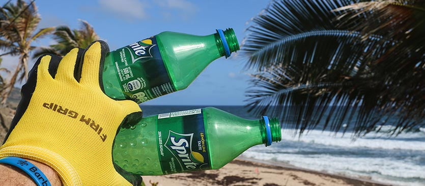 Mann mit gelben Handschuhen hält am Strand zwei Pfandflaschen vor sich